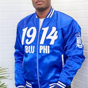 May include: A royal blue satin bomber jacket with white accents. The jacket features the year "1914" and "BLU PHI" in large white letters on the front. A crest is on the left sleeve. The jacket has white stripes on the cuffs and collar.