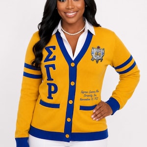 May include: A gold V-neck cardigan with royal blue trim, featuring the Greek letters ΣΓΡ vertically on the right side, a crest on the left chest, and the text Sigma Gamma Rho Sorority, Inc. November 12, 1922 embroidered in blue on the lower left.