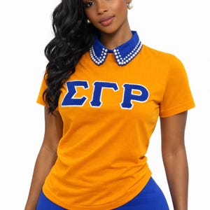 May include: Mustard yellow t-shirt with blue Greek letters and a blue collar adorned with white pearls. The model is wearing blue leggings. The shirt has short sleeves.