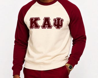 May include: Cream-colored sweatshirt with maroon raglan sleeves and the letters "KAΨ" in maroon. The model is wearing maroon pants and a gold watch. The sweatshirt is a casual style.
