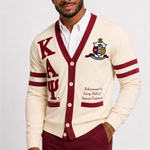 May include: Cream-colored cardigan with burgundy trim, featuring the letters "K A Ψ" and a crest emblem. The text "Achievement in Every Field of Human Endeavor" is displayed on the front. The cardigan has burgundy stripes on the sleeves.