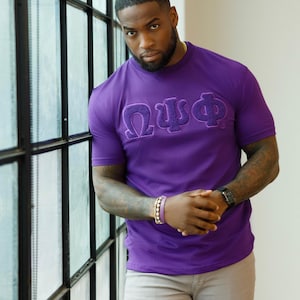 May include: A purple t-shirt with Greek letters embroidered on the front. The shirt is paired with light gray pants. The person is wearing a watch and bracelets. The background includes a window with black frames.