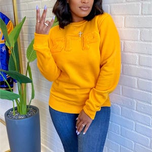 May include: Mustard yellow sweatshirt with raised lettering. The sweatshirt features the Greek letters ΣΓΡ. The model is wearing blue jeans. The background is a white brick wall. A gold necklace with a cross is visible.