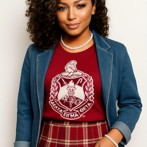 May include: A woman wearing a red t-shirt with a white and red crest, a denim jacket, and a plaid skirt. The t-shirt features Greek letters and a torch design. She is also wearing a pearl necklace and bracelet.