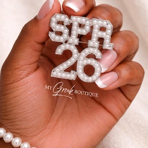 May include: A silver-toned brooch with the letters "SPR" above the number "26", embellished with faux pearls. The text "MY Greek BOUTIQUE" is below the numbers. A pearl bracelet is visible in the image.