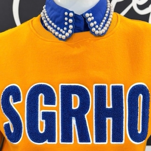May include: Mustard yellow sweatshirt with the word "SGRHO" in blue, outlined in white. A blue collar with pearl embellishments is visible. The sweatshirt is displayed on a mannequin.