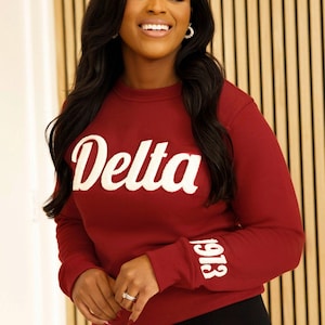 May include: A maroon sweatshirt with the word "Delta" in white script across the chest. The number "1913" is printed in white on the left sleeve. The sweatshirt is paired with black leggings.