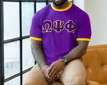 May include: A purple t-shirt with yellow trim and Greek letters is worn with khaki pants. The shirt features the Greek letters "ΩΨΦ" in a contrasting color. The person is seated on a brown leather chair.