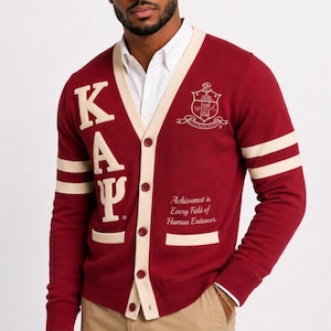 May include: A burgundy cardigan sweater with cream-colored accents, featuring the Greek letters K, A, and Y, along with a crest and the phrase "Achievement in Every Field of Human Endeavor." The sweater has cream-colored stripes on the sleeves.