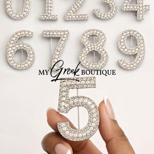May include: A collection of silver-toned rhinestone and pearl-studded number pins, including the number 5 held by a hand. The pins are arranged on a white background. The text "My Greek Boutique" is also visible.