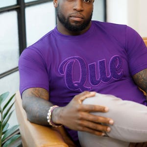 May include: A purple t-shirt with the word "Que" embroidered in a darker purple, worn by a person. The person is seated in a brown leather chair. The person is wearing a bracelet on their wrist.