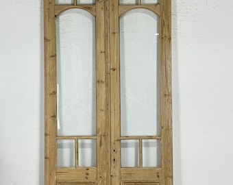 May include: A pair of antique wooden doors with clear glass panels. The doors have a natural wood finish and feature rectangular and arched glass sections. The doors are likely interior doors and are set against a white wall.