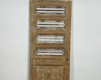 Puede incluir: Una puerta de madera envejecida con un acabado marrón claro. La puerta presenta cuatro paneles rectangulares con listones horizontales y un panel inferior macizo. La puerta está colocada contra una pared blanca y un suelo de color claro.