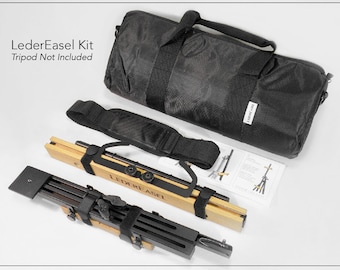 May include: A black canvas bag with a shoulder strap and the text "LederEasel" on the side. The bag contains a wooden and black easel with a strap and the text "LederEasel" on the side. The easel is folded and ready to be assembled. Tripod not included.