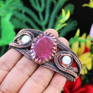 May include: A handcrafted copper wire bracelet featuring a large, faceted, oval-shaped ruby gemstone as the centerpiece. The bracelet also includes two smaller, round, white gemstones and intricate wirework.