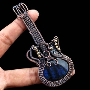 Puede incluir: Un colgante de guitarra envuelto en alambre con una piedra preciosa de labradorita azul. El colgante está hecho de alambre de cobre y presenta un diseño detallado con cuentas doradas.