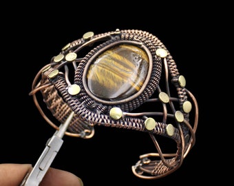 May include: A copper and brass wire wrapped bracelet with a tiger's eye gemstone. The bracelet is open and being held by a pair of pliers.