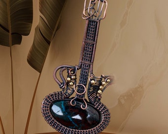 May include: A copper wire guitar pendant featuring a dark blue and green stone inlay. The pendant is decorated with small gold beads.