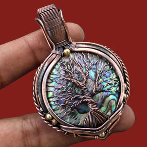May include: Copper wire-wrapped pendant featuring a tree of life design. The pendant has a round, iridescent abalone shell center with a detailed copper wire tree. Accented with small gold-colored beads, the pendant is held in a hand.