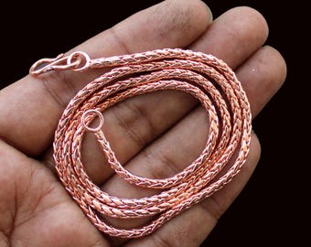 May include: A copper-colored chain necklace with a woven design. The necklace is coiled in a hand, showing the hook clasp and a small ring at the end. The chain has a shiny, metallic finish.