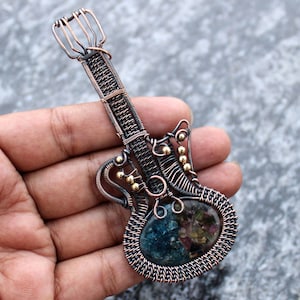 Könnte beinhalten: Ein drahtgewickelter Gitarrenanhänger mit einem blauen und grünen Edelstein in der Mitte. Die Gitarre ist aus Kupferdraht gefertigt und hat goldene Perlen am Kopf und am Korpus.