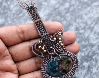Puede incluir: Un colgante de guitarra envuelto en alambre con una piedra preciosa azul y verde en el centro. La guitarra está hecha de alambre de cobre y tiene cuentas doradas en la pala y el cuerpo.