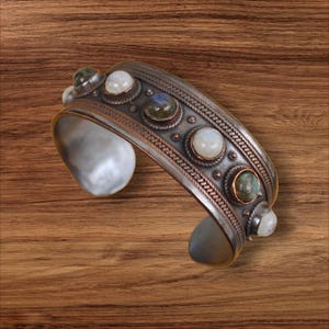 May include: A silver cuff bracelet with a decorative design and inlaid gemstones. The bracelet features a series of round, bezel-set stones in shades of white, green, and blue. The metal has a textured, antique finish.