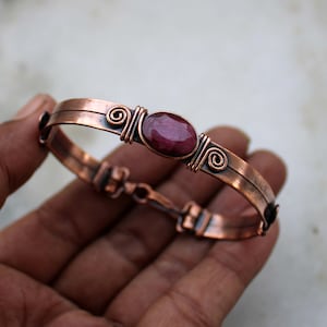 May include: A copper bracelet featuring a central oval-shaped faceted dark pink gemstone. The metal band displays a weathered finish with decorative wire-wrapped spiral accents on either side of the stone and a hinged clasp mechanism.