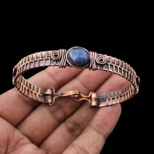 May include: Copper wire bracelet with a woven design and a central blue stone. The bracelet has a hook clasp and decorative wire detailing around the stone. The copper has a warm, reddish-brown hue.