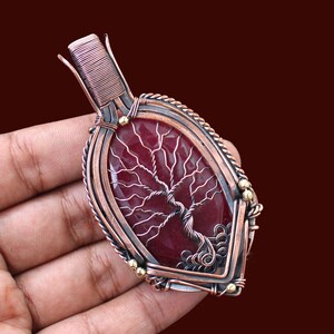 May include: A handcrafted pendant featuring a red gemstone with a wire tree design. The pendant is encased in copper wire with decorative accents. The overall shape is an elongated oval, perfect for a necklace.