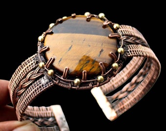 May include: A copper wire wrapped bracelet with a large round tiger's eye gemstone in the center. The bracelet is made of woven copper wire and has a clasp closure.