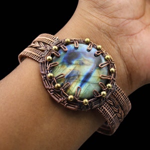 Op de afbeelding: Een koperkleurige manchetarmband met een grote, ovale labradorietsteen met blauwe, groene en gele iriserende kleuren. De steen is omcirkeld door een decoratief draadontwerp met kleine goudkleurige kralen. De armband heeft een geweven band.