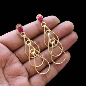 Peut inclure: Boucles d'oreilles pendantes dorées avec une pierre ovale rouge rubis en haut. Les boucles d'oreilles présentent un design en goutte d'eau ouvert et superposé. Le fond est noir.