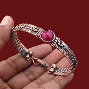 May include: A handcrafted copper bracelet with a woven design and a central, round, red gemstone. The bracelet features intricate wirework and decorative swirls. The clasp is a hook and eye style.