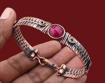 May include: A handcrafted copper bracelet with a woven design and a central, round, red gemstone. The bracelet features intricate wirework and decorative swirls. The clasp is a hook and eye style.