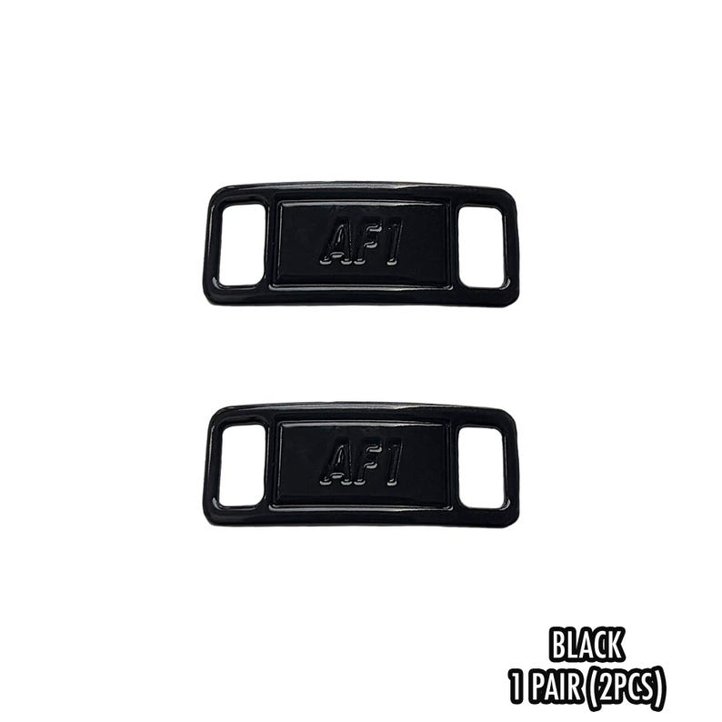 May include: Two black rectangular shoe accessories with the text "AF1" embossed on each. Each accessory has two rectangular holes. The text "BLACK 1 PAIR (2PCS)" is at the bottom.