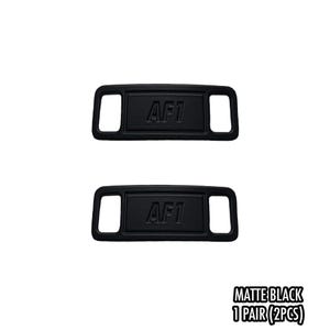 May include: Two matte black shoe lace tags with the text "AF1" embossed on each. The tags are rectangular with a curved top and bottom and a rectangular hole on each side. The text "MATTE BLACK 1 PAIR (2PCS)" is at the bottom.