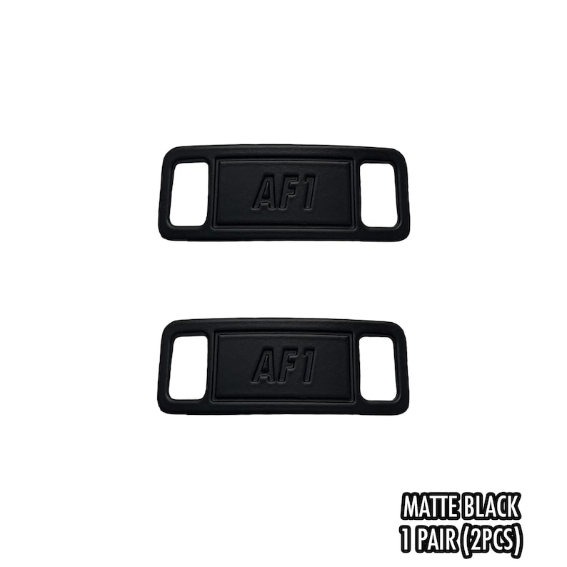 May include: Two matte black shoe lace tags with the text "AF1" embossed on each. The tags are rectangular with a curved top and bottom and a rectangular hole on each side. The text "MATTE BLACK 1 PAIR (2PCS)" is at the bottom.