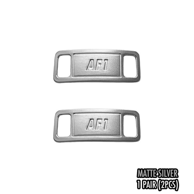 May include: Two matte silver shoe lace tags with the text "AF1" embossed on each. The tags have a rectangular shape with a slot on each end. The bottom right corner of the image has the text "MATTE SILVER 1 PAIR (2PCS)".