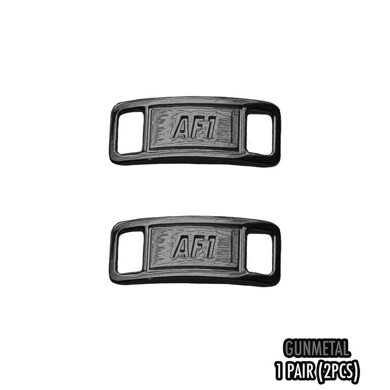 May include: Two gunmetal-colored shoe lace tags with the text "AF1" embossed on each. The tags have a rectangular shape with rounded corners and two rectangular openings. The text "GUNMETAL 1 PAIR (2PCS)" is also visible.