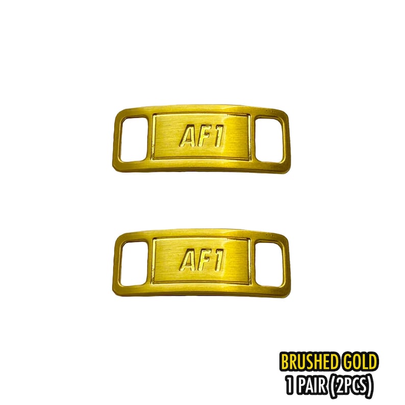 May include: Two brushed gold metal shoe lace tags with the text "AF1" on each. The tags have a rectangular shape with rounded corners and two rectangular holes on each end. The text "BRUSHED GOLD 1 PAIR (2PCS)" is in the bottom right corner.
