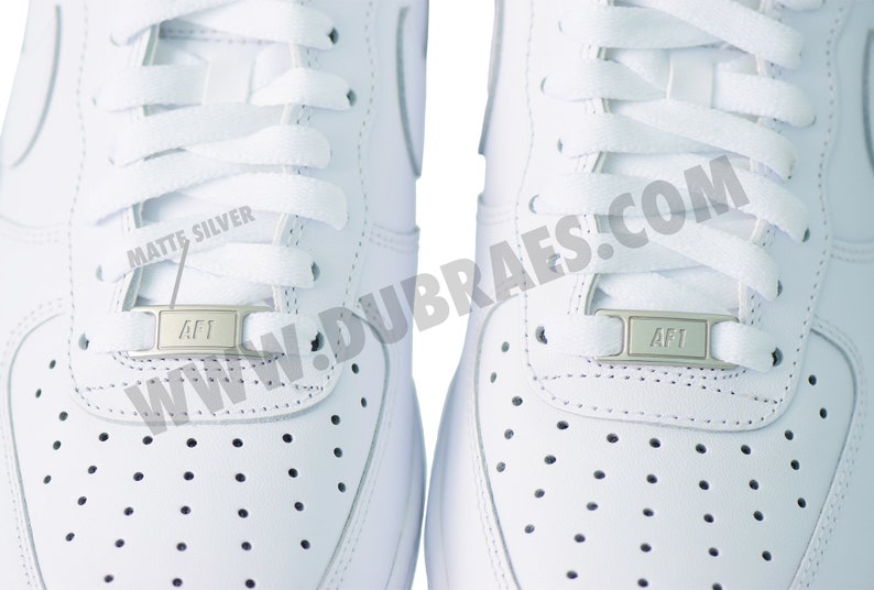 May include: Close-up of two white sneakers with white laces. Each shoe has a silver AF1 lace lock. The shoes have small perforations on the toe box and are made of a smooth, white material.