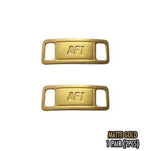 May include: Two matte gold shoe lace tags with the text "AF1" embossed on each. The tags are rectangular with rounded corners and a slot on each end for the laces. The text "MATTE GOLD 1 PAIR (2PCS)" is printed in black.