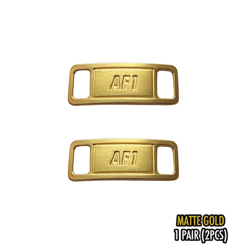 May include: Two matte gold shoe lace tags with the text "AF1" embossed on each. The tags are rectangular with rounded corners and a slot on each end for the laces. The text "MATTE GOLD 1 PAIR (2PCS)" is printed in black.