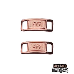 May include: Two rose gold AF1 shoe lace tags. Each tag is rectangular with rounded corners and the letters "AF1" engraved on the front. The tags have openings on each end for the laces. The text "ROSE GOLD 1 PAIR (2PCS)" is at the bottom.