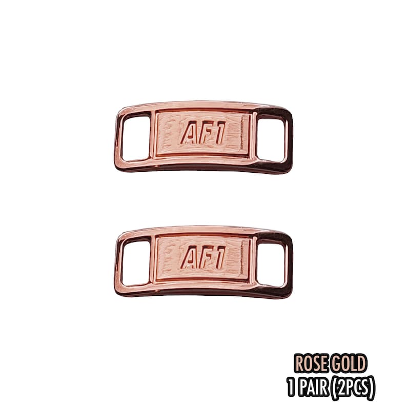May include: Two rose gold AF1 shoe lace tags. Each tag is rectangular with rounded corners and the letters "AF1" engraved on the front. The tags have openings on each end for the laces. The text "ROSE GOLD 1 PAIR (2PCS)" is at the bottom.