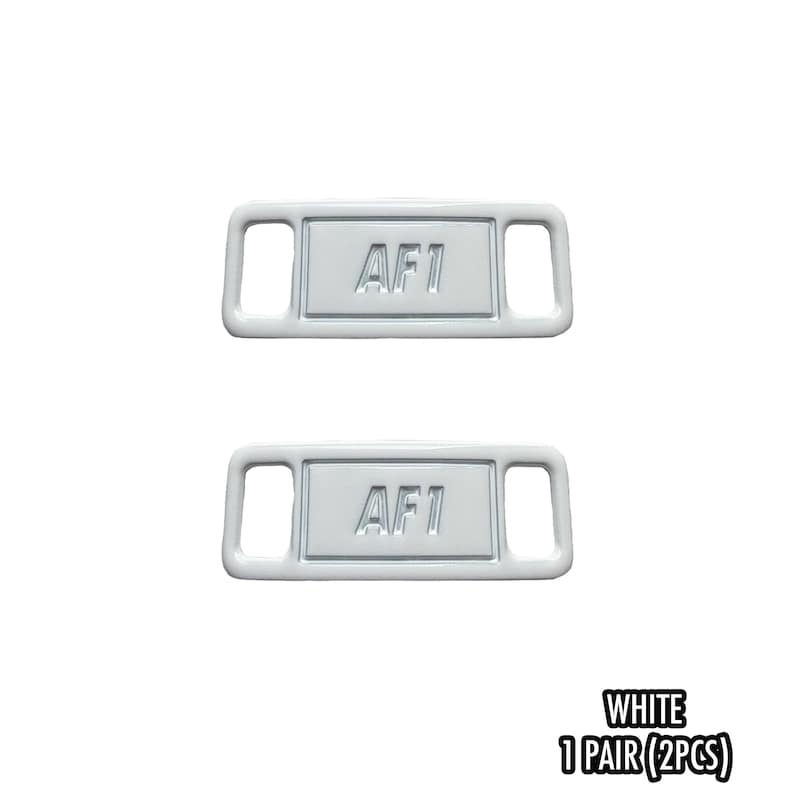 May include: Two white shoe lace tags with the text "AF1" in the center. The tags have a rectangular shape with rounded corners and two rectangular holes on each side. The text "WHITE 1 PAIR (2PCS)" is at the bottom.