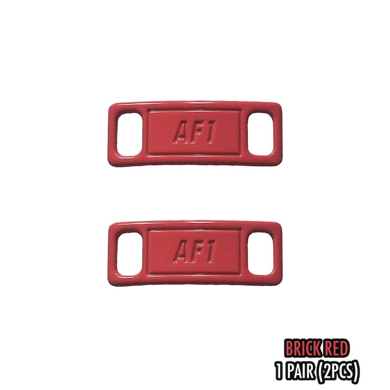 May include: Two brick red shoe tags with the text "AF1" embossed on each. The tags have a rectangular shape with rounded corners and two holes for laces. The bottom right corner of the image has the text "BRICK RED 1 PAIR (2PCS)".