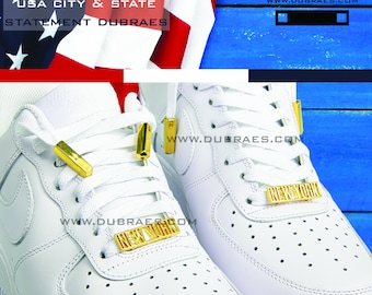 May include: A pair of white sneakers with gold lace locks and a gold "NEW YORK" charm on each shoe. The shoes are on a blue wooden background.