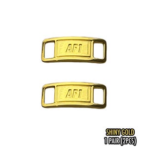 May include: Two shiny gold AF1 shoe lace tags. Each tag is rectangular with rounded corners and a rectangular opening on each end. The text "AF1" is embossed on the front. The text "SHINY GOLD 1 PAIR (2PCS)" is in the bottom right corner.
